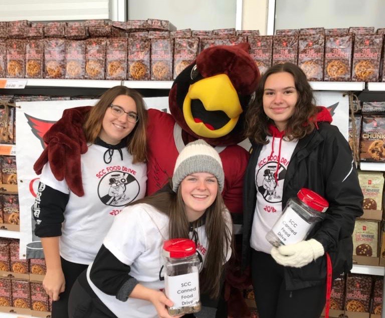 Three students from St. Charles College stand with the school mascot alongside piles of canned food donations